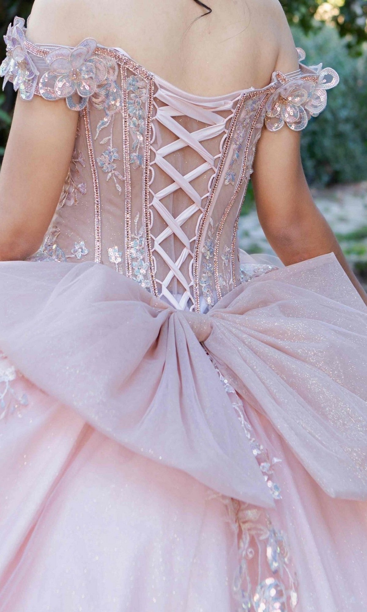 A person is photographed from behind outdoors wearing the Quinceanera Dress 8370J by Cinderella Couture, featuring an embellished pink corset bodice, floral appliqués, and a large tulle bow.